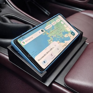 Op de afbeelding: Een smartphone met blauwe hoes toont een navigatiekaart en ligt op een zwarte telefoonhouder. De telefoon bevindt zich in een auto met bordeauxrode leren stoelen en een donker dashboard.