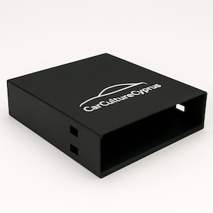 Puede incluir: Una caja rectangular negra con bordes redondeados. La superficie superior presenta el texto blanco "CarCultureCyprus" con un gráfico de coche estilizado. La caja tiene dos pequeños recortes rectangulares en un lado y uno más grande en el otro.