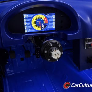 Può includere: Interni blu di un'auto da corsa, con cruscotto e volante in evidenza. Il display digitale mostra indicatori. Il volante è nero. Sono visibili interruttori e spie.