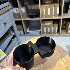 May include: A black dual cup holder accessory featuring two circular openings connected by a central bridge. The object has a smooth, matte finish and a structured, rigid design with vertical lines visible along the interior walls of the cups.