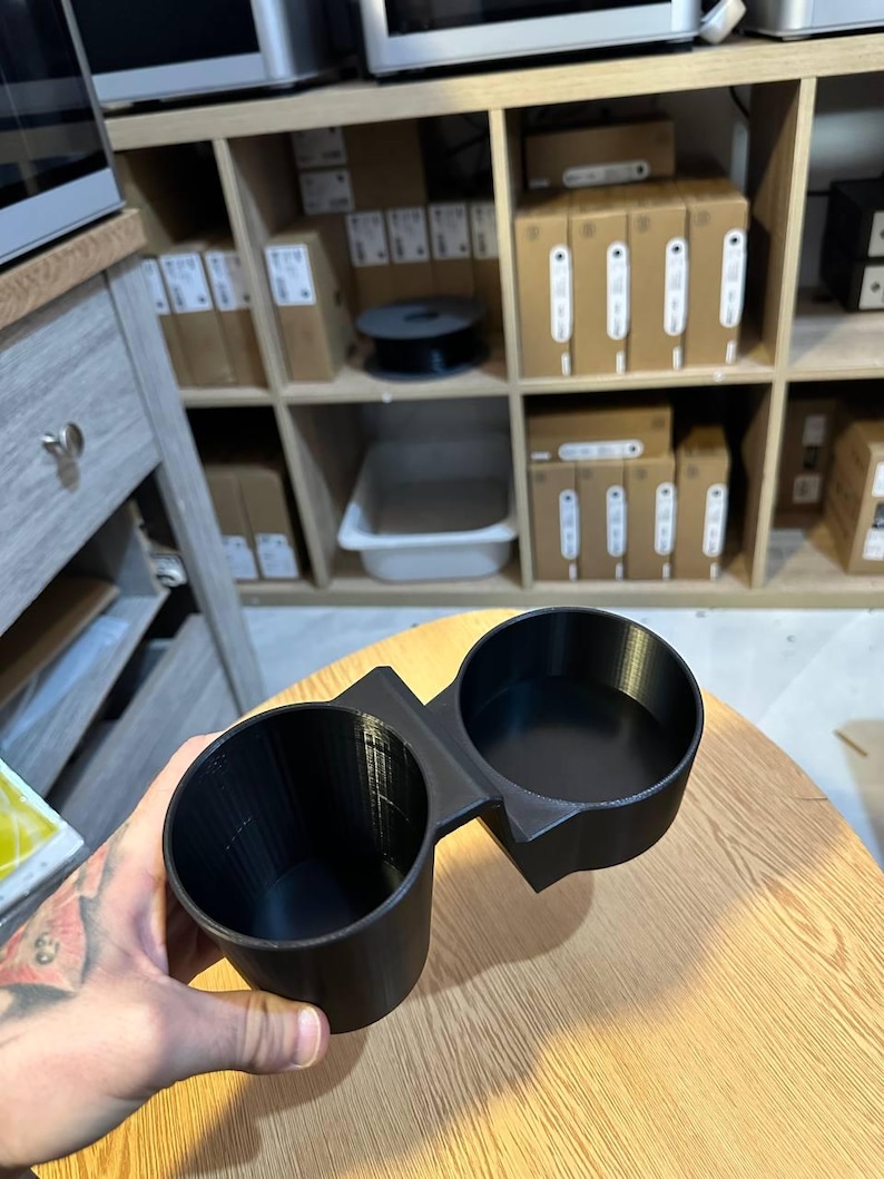 May include: A black dual cup holder accessory featuring two circular openings connected by a central bridge. The object has a smooth, matte finish and a structured, rigid design with vertical lines visible along the interior walls of the cups.