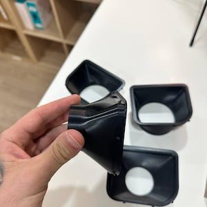 May include: Three black, funnel-shaped plastic components with a square-like opening at one end and a circular opening at the other. The objects feature a tapered, smooth design with a rigid structure, designed for directing airflow or light.