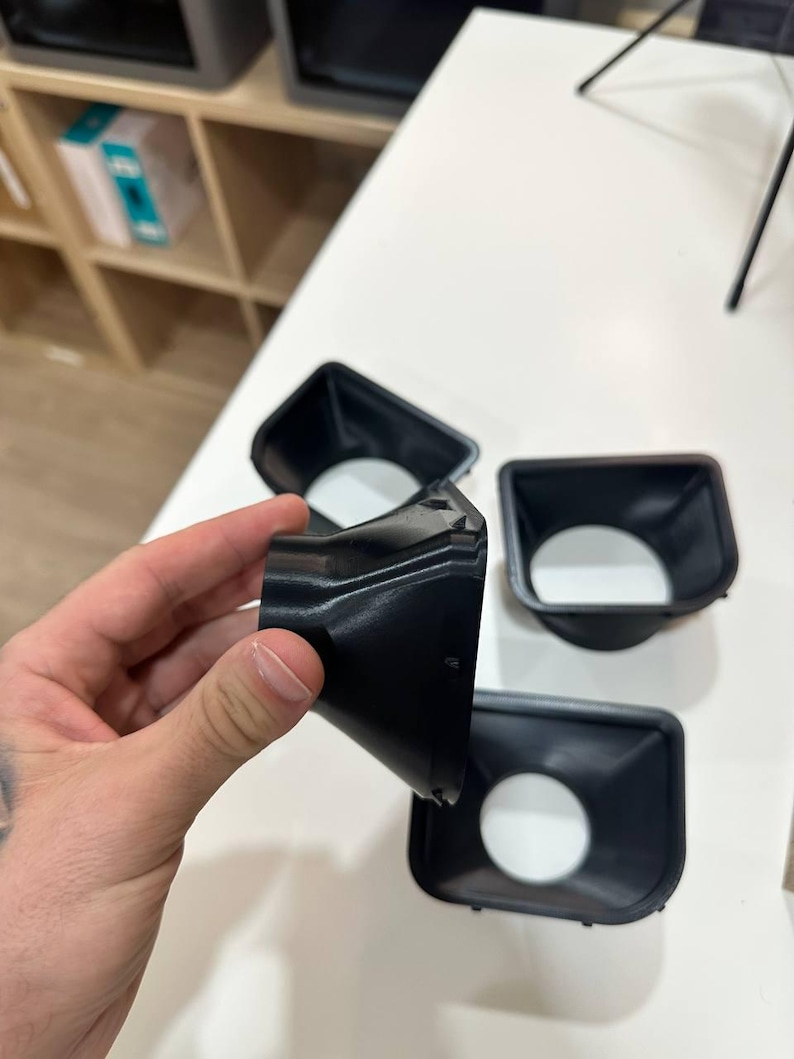 May include: Three black, funnel-shaped plastic components with a square-like opening at one end and a circular opening at the other. The objects feature a tapered, smooth design with a rigid structure, designed for directing airflow or light.