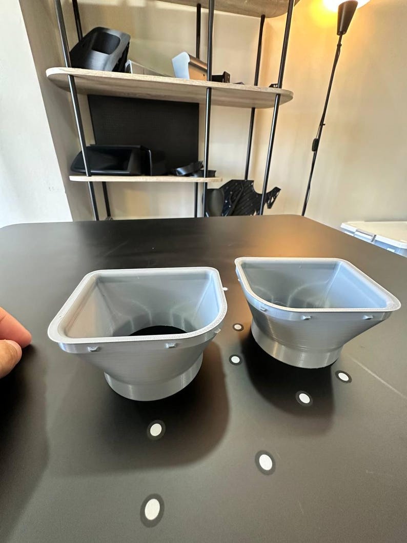 May include: Two light gray, funnel-shaped plastic components with a wide, square-like opening that tapers down to a circular base. Each piece features a flat, wide rim with small, evenly spaced protrusions along the outer edge of the top opening.