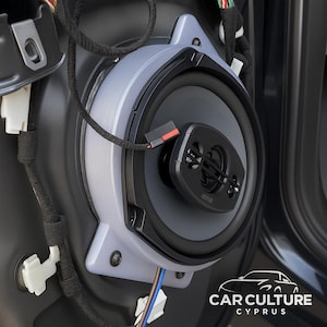May include: A car audio speaker system in grey and black, mounted within a car door panel. The speaker features a central tweeter and a larger woofer, with visible wiring and connectors. The design is modern and sleek.