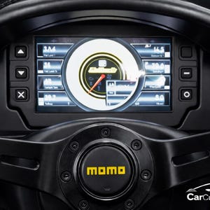 Può includere: Primo piano di un volante nero per auto con cruscotto digitale. Il cruscotto mostra vari indicatori e dati, tra cui velocità e prestazioni del motore. Il centro del volante presenta il logo giallo "momo".