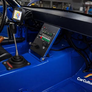 Può includere: Vista interna di un'auto da corsa blu. Sono presenti un volante nero, un display digitale e un pannello di interruttori. Sono visibili anche una leva del cambio e il logo CarCultureCyprus.
