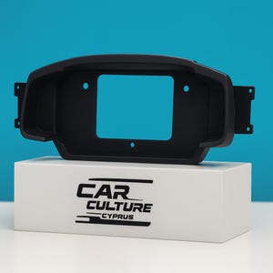 May include: A black car dashboard bezel with a rectangular opening, mounted on a white box. The bezel has mounting tabs and several holes. The white box has the text "CAR CULTURE CYPRUS" printed on it in black.