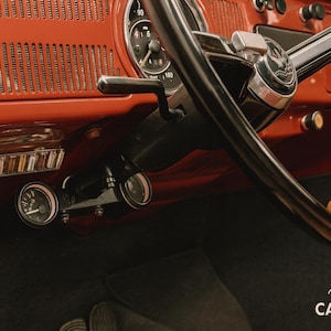 May include: The interior of a vintage vehicle featuring a bright orange dashboard with a black steering wheel, a circular speedometer, and two small auxiliary gauges mounted below the steering column. A wooden gear shift knob is visible on the right.
