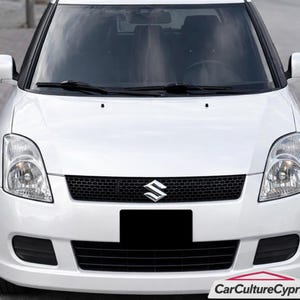 May include: A white Suzuki Swift car with a black grille and headlights. The Suzuki logo is centered on the grille. The car has a black license plate and the CarCultureCyprus logo in the bottom right corner.