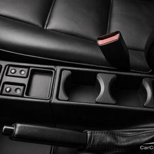 Pode incluir: Uma consola central automóvel preta com um suporte duplo para copos, um pequeno compartimento de arrumação e um painel de controlo com interruptores de vidros e um botão de luzes de emergência. Uma alavanca de travão de mão é visível.