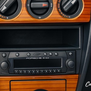 以下が含まれることがあります： ポルシェCD 10-21ラジオを備えた車のダッシュボードのクローズアップ。 ダッシュボードは木目調仕上げで、黒いコントロールとデジタルディスプレイを備えています。 画像には、通気孔と収納コンパートメントも表示されています。