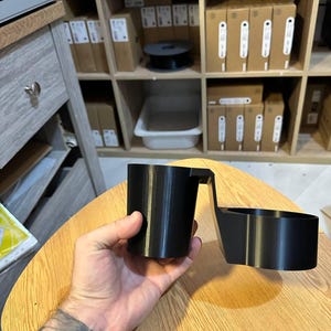 May include: A black 3D-printed plastic cup holder attachment featuring a cylindrical cup-shaped section connected by a horizontal bridge to a secondary circular ring-shaped holder, designed for mounting onto a flat surface edge.