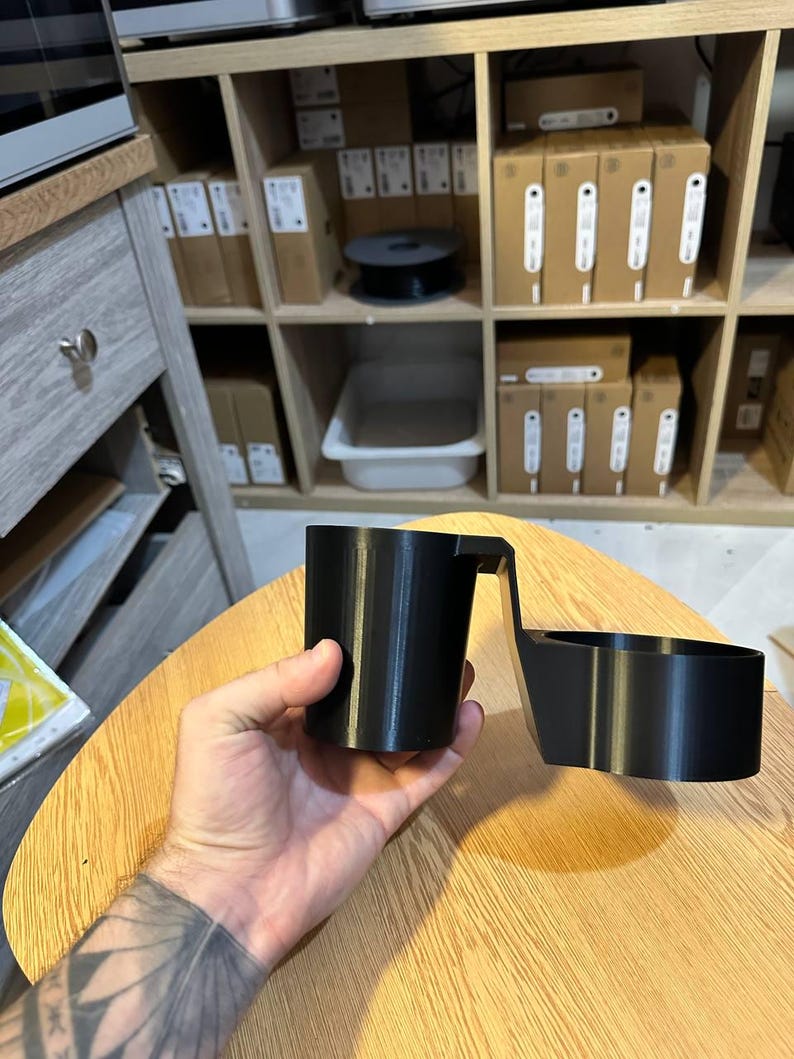 May include: A black 3D-printed plastic cup holder attachment featuring a cylindrical cup-shaped section connected by a horizontal bridge to a secondary circular ring-shaped holder, designed for mounting onto a flat surface edge.