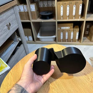 May include: A matte black 3D printed dual cup holder accessory featuring two cylindrical compartments of different sizes connected by a rigid angular bridge, designed to hold beverage containers securely in a vehicle or workspace.