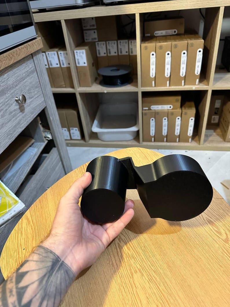 May include: A matte black 3D printed dual cup holder accessory featuring two cylindrical compartments of different sizes connected by a rigid angular bridge, designed to hold beverage containers securely in a vehicle or workspace.