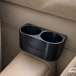 Puede incluir: Portavasos negro para coche con dos aberturas circulares, situado entre los asientos del coche. La palabra "Mazda" está grabada en la parte delantera. El portavasos es de material negro mate.