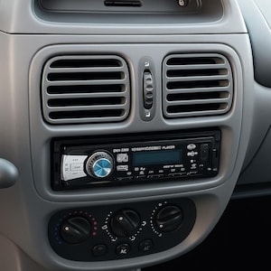 May include: A close-up of a car's dashboard, featuring a gray plastic design. The dashboard includes air vents, a digital radio with a blue dial, and climate control knobs. A gear shift is visible at the bottom.