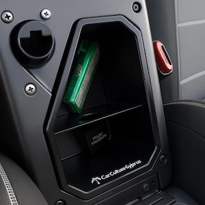 Op de afbeelding: Een zwart opbergvak voor de middenconsole van een auto met een zeshoekig frame, een horizontale plank en een ronde uitsparing. Het interieur bevat een groen rechthoekig object, een kleine zwarte inzet met het label Magic Mealmate en de tekst CarCultureGyprus.