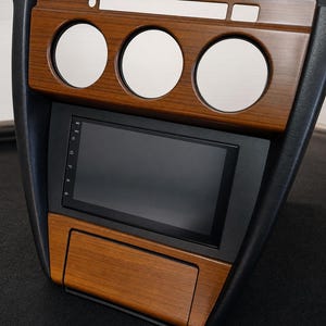 Può includere: Un pannello per console centrale di un cruscotto auto con finitura effetto legno, dotato di tre aperture circolari per indicatori nella parte superiore, uno schermo digitale nero rettangolare e un vano portaoggetti inferiore.