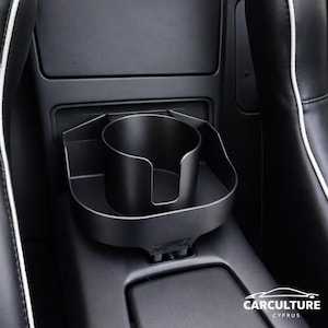 May include: A black car cup holder with a circular opening and a U-shaped cutout for easy access. The cup holder is designed to fit into a car's center console. The image shows the cup holder installed in a car interior.