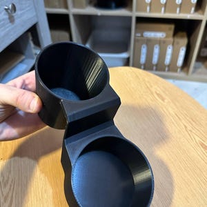 May include: A matte black 3D printed dual cup holder featuring two circular compartments connected by a central bridge. The object has a smooth, uniform finish with visible layer lines from the manufacturing process and a sturdy, rigid structure.