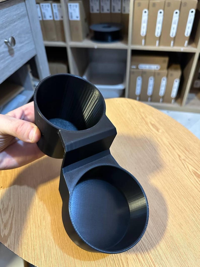May include: A matte black 3D printed dual cup holder featuring two circular compartments connected by a central bridge. The object has a smooth, uniform finish with visible layer lines from the manufacturing process and a sturdy, rigid structure.