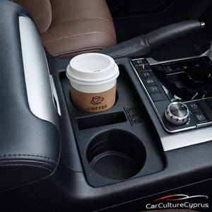May include: A disposable coffee cup with a white lid sits in a car's cup holder. The cup has a brown sleeve with the word "COFFEE" and a coffee cup graphic. The interior of the car features brown leather seats and black trim.