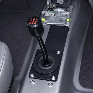 May include: A black gear shift lever with red markings for the gears, mounted on a black plate. The plate is secured with silver bolts to a gray console. The image is of a car interior.