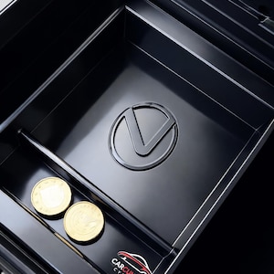 Könnte beinhalten: Eine schwarze Auto-Konsolen-Ablageschale mit silbernem Emblem und zwei Goldmünzen. Die Schale hat einen geteilten Bereich und ein Logo "CARCULTURE CYPRUS".