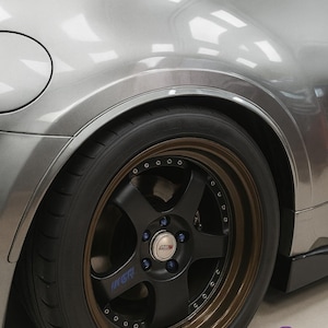 Pode incluir: Close-up da roda traseira de um carro desportivo prateado. O pneu preto está montado numa jante de liga leve preta e bronze com porcas azuis. A jante tem o texto "INGER" em azul. A carroçaria do carro é prateada.