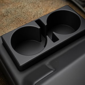 May include: A black plastic car console cup holder with two circular openings. The cup holder is rectangular and sits on top of a larger, textured black surface. The background is a tan, woven material.