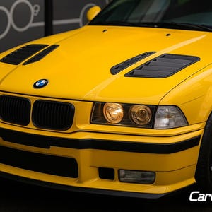 May include: A vibrant yellow BMW car with black detailing on the front. Features include black vents on the bonnet, a black front grill, and black wheels. The headlights are illuminated, and the BMW emblem is visible. The image is a close-up.