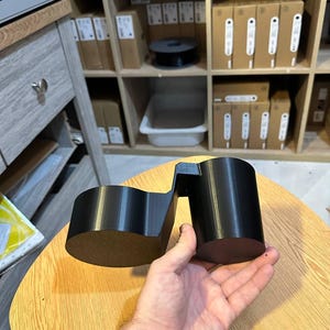 May include: A black 3D printed dual cup holder accessory featuring two cylindrical compartments connected by a central bridge, designed to hold two beverage containers simultaneously with a smooth, matte finish and rigid construction.