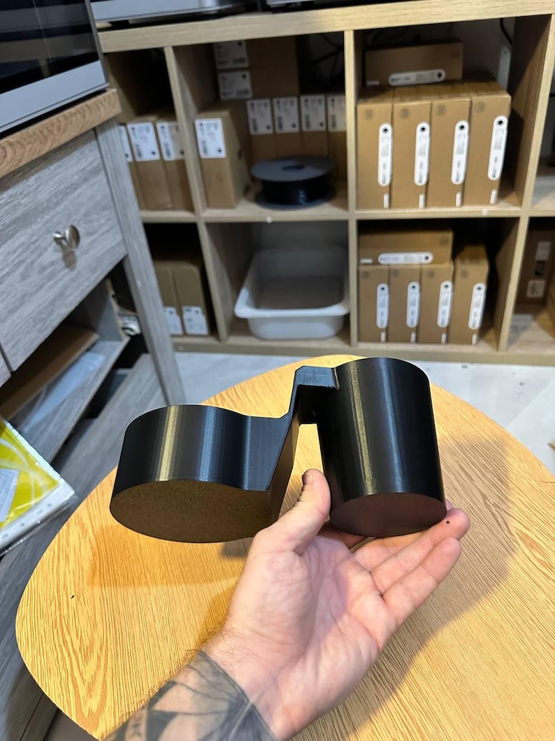 May include: A black 3D printed dual cup holder accessory featuring two cylindrical compartments connected by a central bridge, designed to hold two beverage containers simultaneously with a smooth, matte finish and rigid construction.