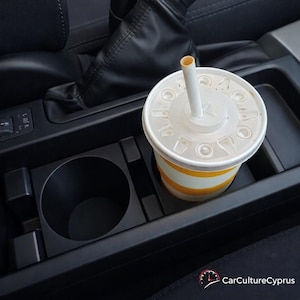 May include: A fast-food beverage cup with a white lid and straw sits in a car's cup holder. The cup has yellow and white stripes. The car's interior is black.