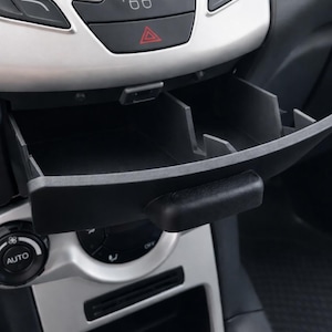 Może przedstawiać: Zbliżenie na deskę rozdzielczą samochodu, z naciskiem na konsolę środkową. Konsola ma srebrno-czarną konstrukcję z przyciskami do różnych funkcji. Widoczny jest otwarty czarny schowek.