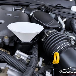 May include: A white plastic funnel inserted into a black automotive oil filler neck within a car engine bay. The engine compartment features various black rubber hoses, metallic components, and a bright yellow oil dipstick handle visible nearby.