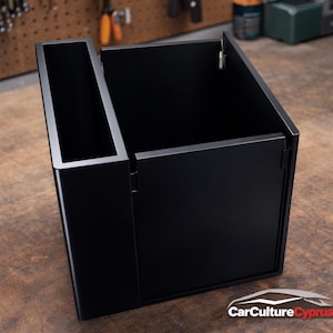 May include: A black storage container with a compartment and a separate section. The container has a hinged door and is made of a matte black material. The container is on a wooden surface.