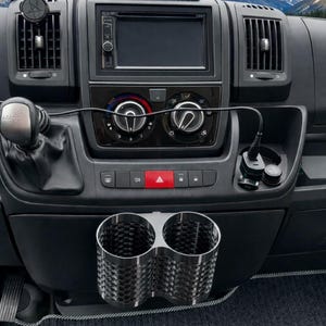 Può includere: Cruscotto nero del veicolo con sistema di infotainment integrato, bocchette dell'aria e portabicchieri doppio. Un piccolo schermo è montato sopra il cruscotto. Lo sfondo mostra una catena montuosa.