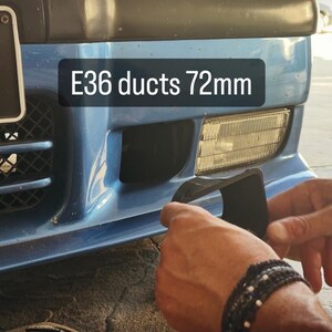 May include: A black plastic brake cooling duct component for an E36 vehicle, labeled E36 ducts 72mm, positioned near the front bumper of a blue car with a rectangular fog light and a black grille with a mesh pattern visible on the left side.