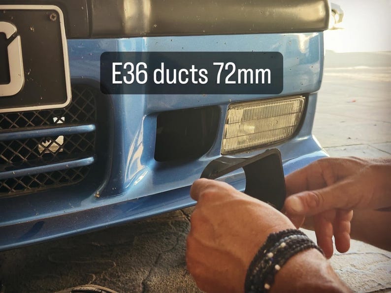 May include: A black plastic brake cooling duct component for an E36 vehicle, labeled E36 ducts 72mm, positioned near the front bumper of a blue car with a rectangular fog light and a black grille with a mesh pattern visible on the left side.