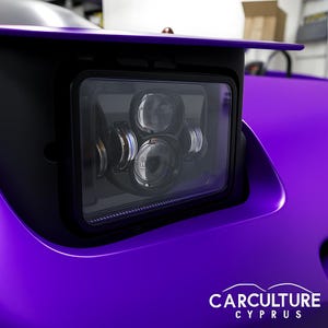 May include: Close-up of a black rectangular LED headlight with multiple circular lights. The headlight is set against a vibrant purple car body. The image includes the text "CARCULTURE CYPRUS" in white.