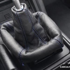 Puede incluir: Fuelle de palanca de cambios de cuero negro con costuras azules, que se muestra en el interior de un coche. El fuelle envuelve la palanca de cambios y forma parte del conjunto del cambio de marchas del coche.