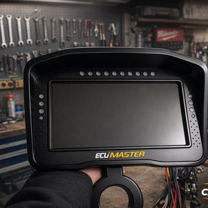 Op de afbeelding: Zwart digitaal ECU Master display met een groot scherm en meerdere indicatielampjes. De tekst "ECU MASTER" is geel. De achtergrond toont een werkplaats met gereedschap en apparatuur.