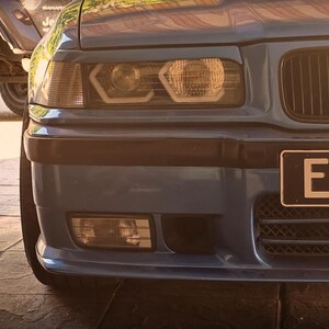 May include: A blue car front end featuring a black grille, rectangular headlights with integrated circular light elements, and a black bumper. A partial license plate with the letters EN is visible on the right side of the front bumper area.