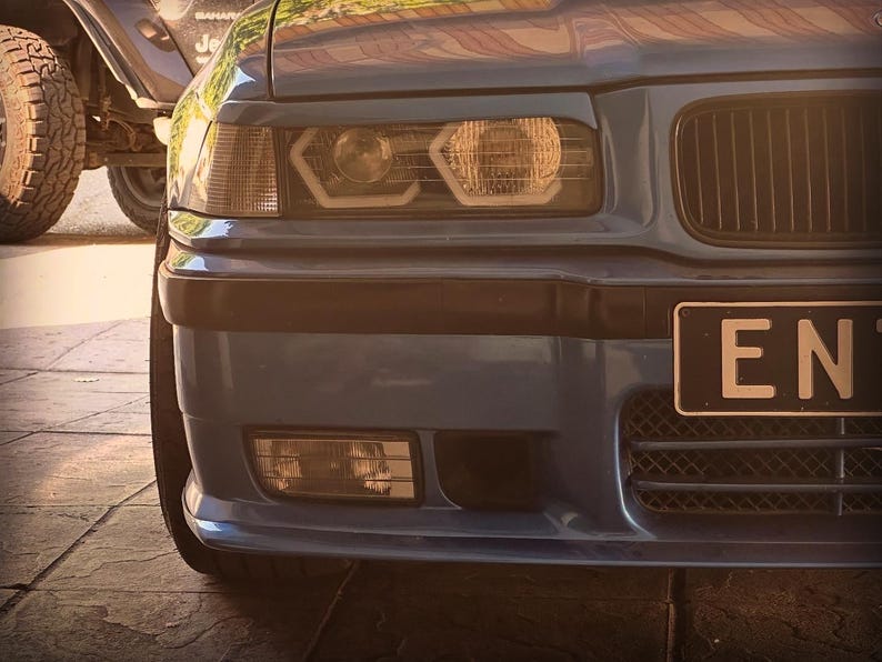 May include: A blue car front end featuring a black grille, rectangular headlights with integrated circular light elements, and a black bumper. A partial license plate with the letters EN is visible on the right side of the front bumper area.