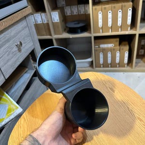 May include: A black, 3D-printed dual cup holder featuring two circular, open-top compartments connected by a central bridge. The object has a matte finish with visible layer lines from the manufacturing process and a smooth, uniform texture.