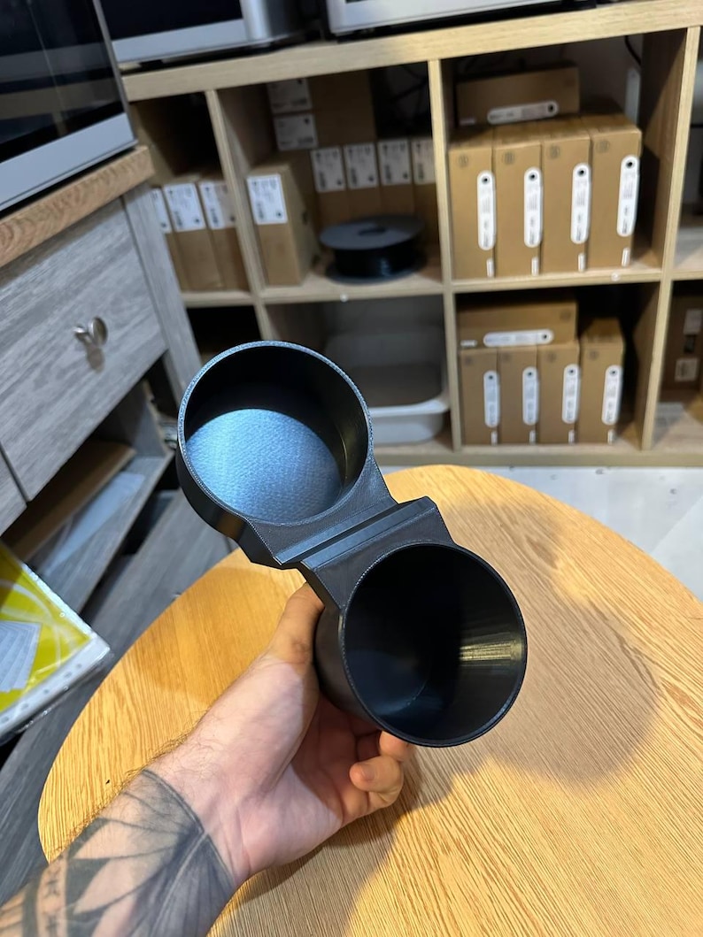 May include: A black, 3D-printed dual cup holder featuring two circular, open-top compartments connected by a central bridge. The object has a matte finish with visible layer lines from the manufacturing process and a smooth, uniform texture.