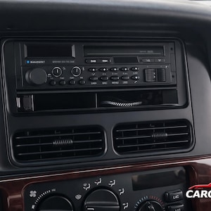 Könnte beinhalten: Nahaufnahme eines Armaturenbretts mit einem Blaupunkt-Autoradio. Das Radio verfügt über einen Kassettenrekorder und ein digitales Display. Unter dem Radio befinden sich Lüftungsschlitze und Klimaanlagenknöpfe. Das Armaturenbrett ist schwarz mit Holzmaserungsakzenten.